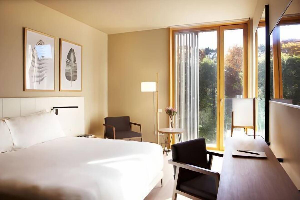 A hotel room with a large bed, a desk, a chair, a small table with a vase, and framed artwork on the wall.