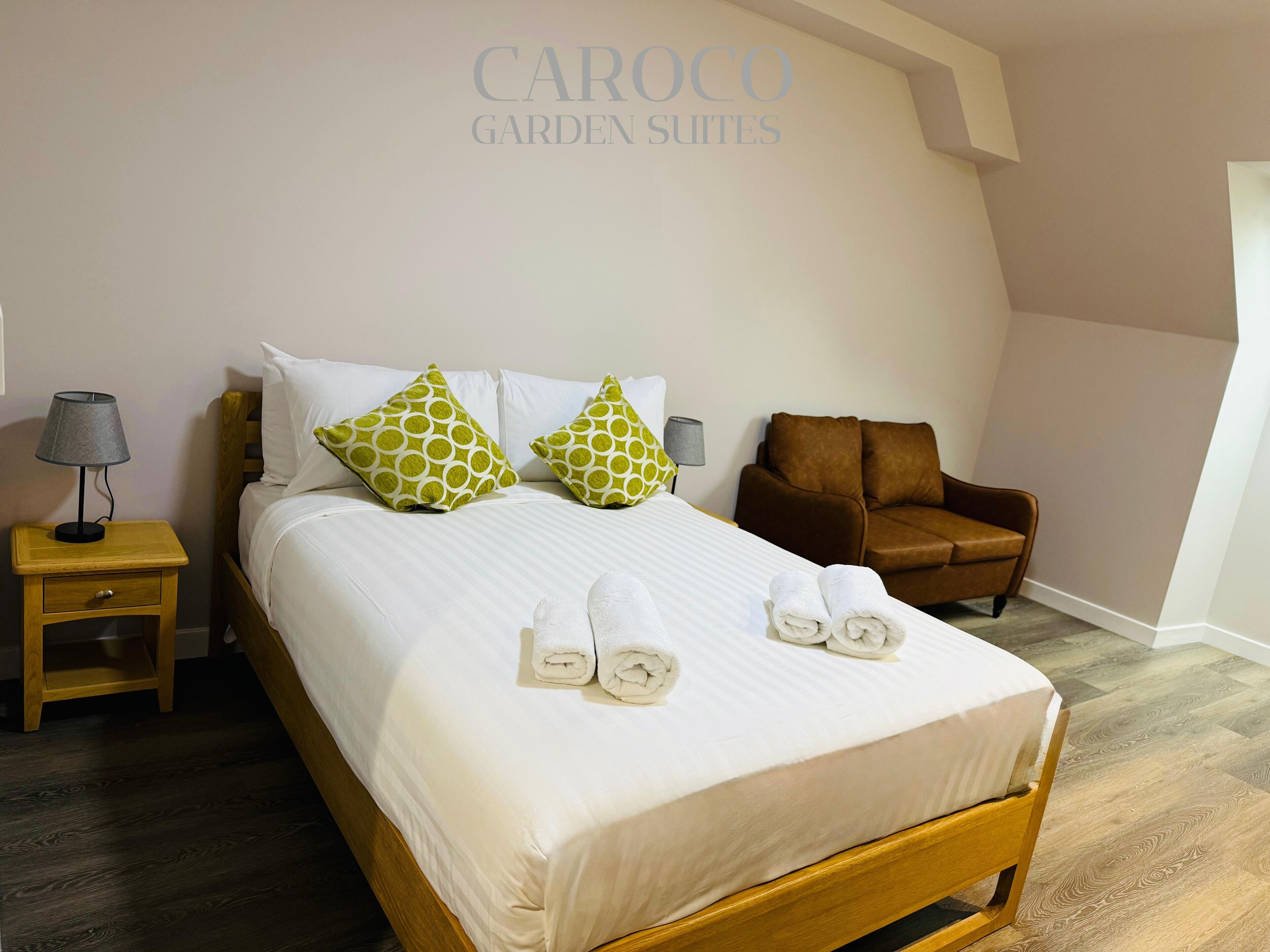 Una cama bien hecha con sábanas blancas y dos almohadas estampadas en verde, un sillón de cuero marrón, una mesita de noche de madera con una lámpara, y un cartel en la pared que dice 'CAROCO GARDEN SUITES'.