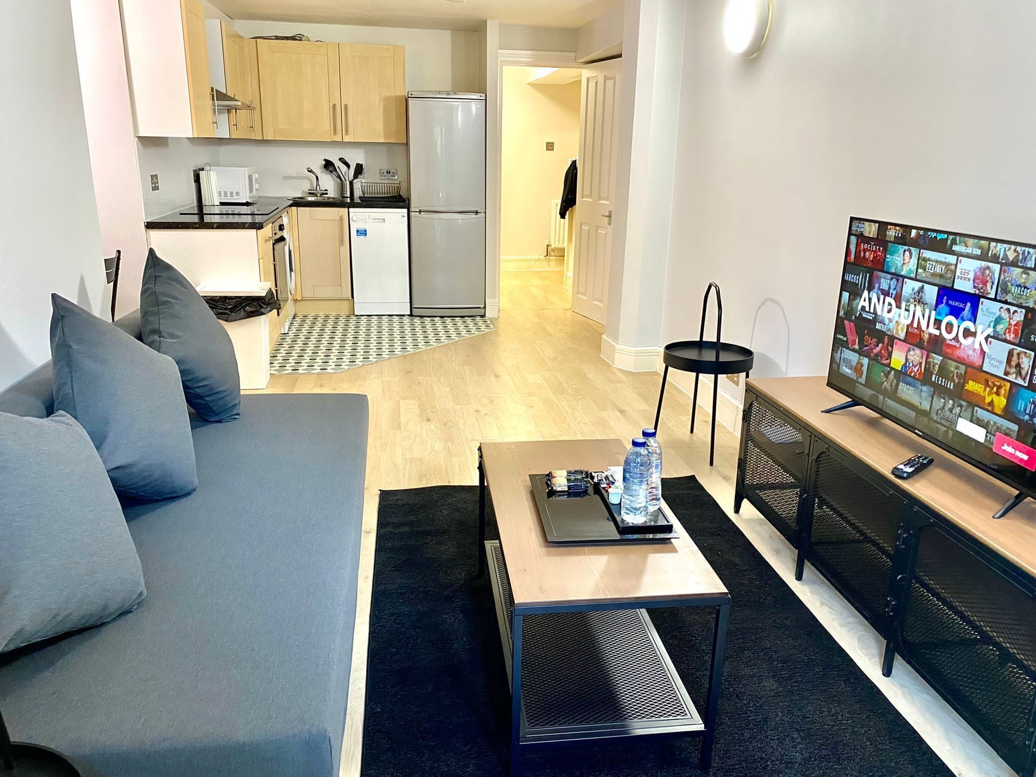 A modern living room with a grey sofa, a coffee table, a TV, and a kitchen in the background.
