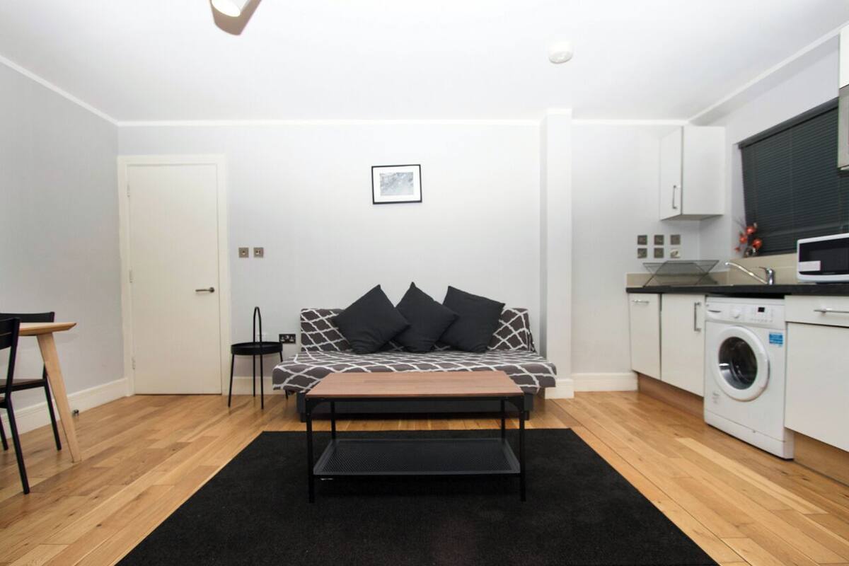 Ett modernt och kompakt bostadsutrymme med en soffa, ett soffbord, en tvättmaskin och ett litet köksområde.