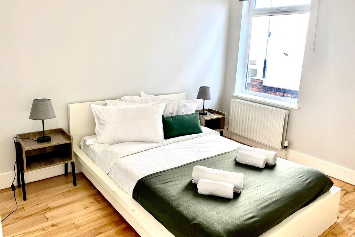 En snyggt bäddad säng med vita sängkläder och en grön pläd, två sänglampor, ett träbord och ett fönster med utsikt över en byggnad.
