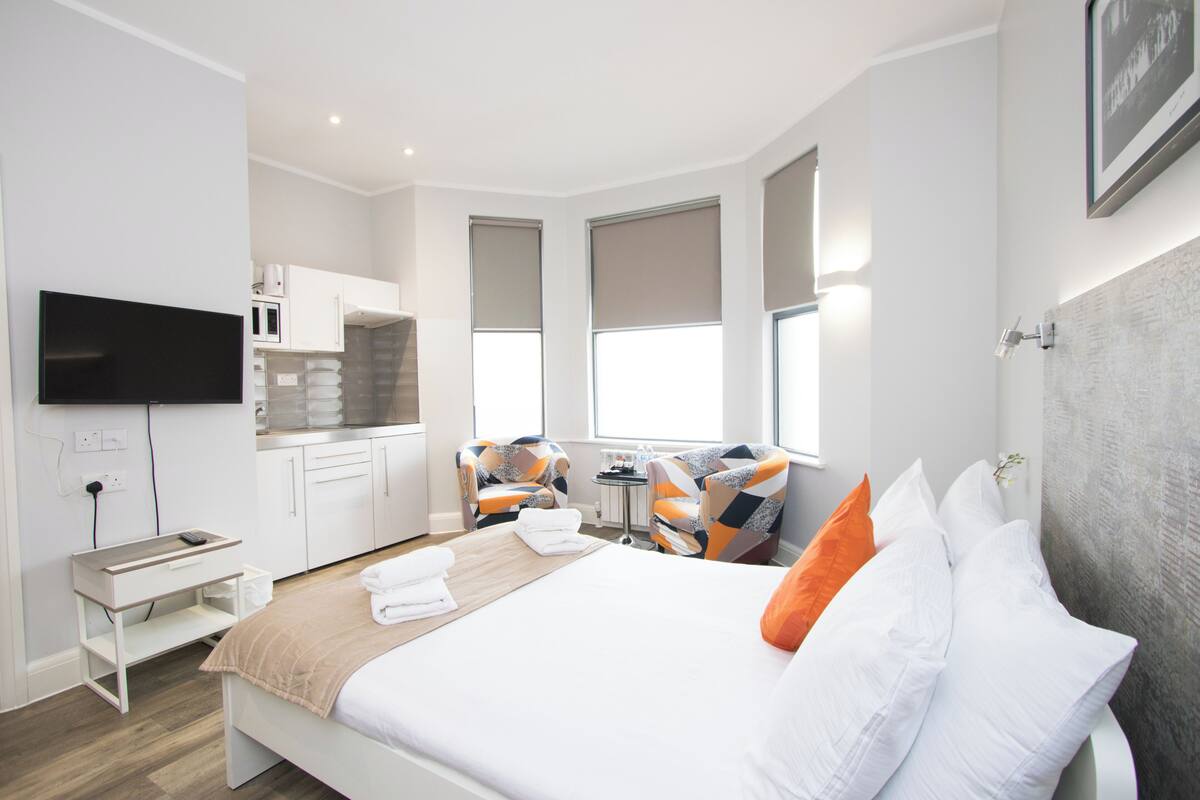 Ett modernt hotellrum med en säng, ett skrivbord, en stol, en TV och ett litet kök.