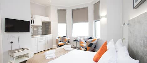 A modern hotel room with a bed, a desk, a chair, a TV, and a kitchenette.