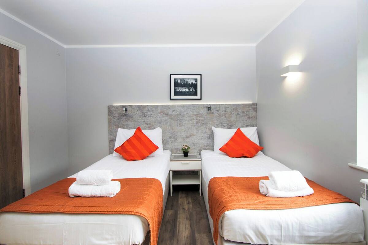 Ett hotellrum med två separata sängar, var och en med ett orangefärgat överkast och en vit kudde. Mellan sängarna står ett litet bord med en vas med blommor.