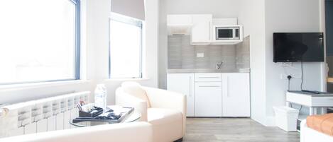 Un espace de vie moderne comprenant un canapé, une table à manger, un téléviseur et un coin cuisine avec des meubles blancs et un four à micro-ondes.