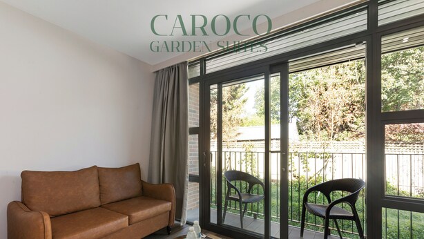 A living room with a brown sofa, a small round table with a tray, and two chairs on a balcony with a view of greenery. - Westciti Caroco Aparthotel (Croydon)