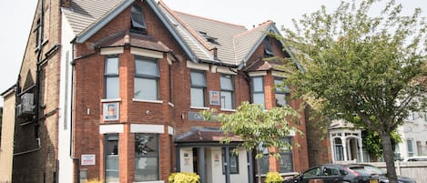 A two-story brick building with a corner entrance, a small garden, and a parked car.
