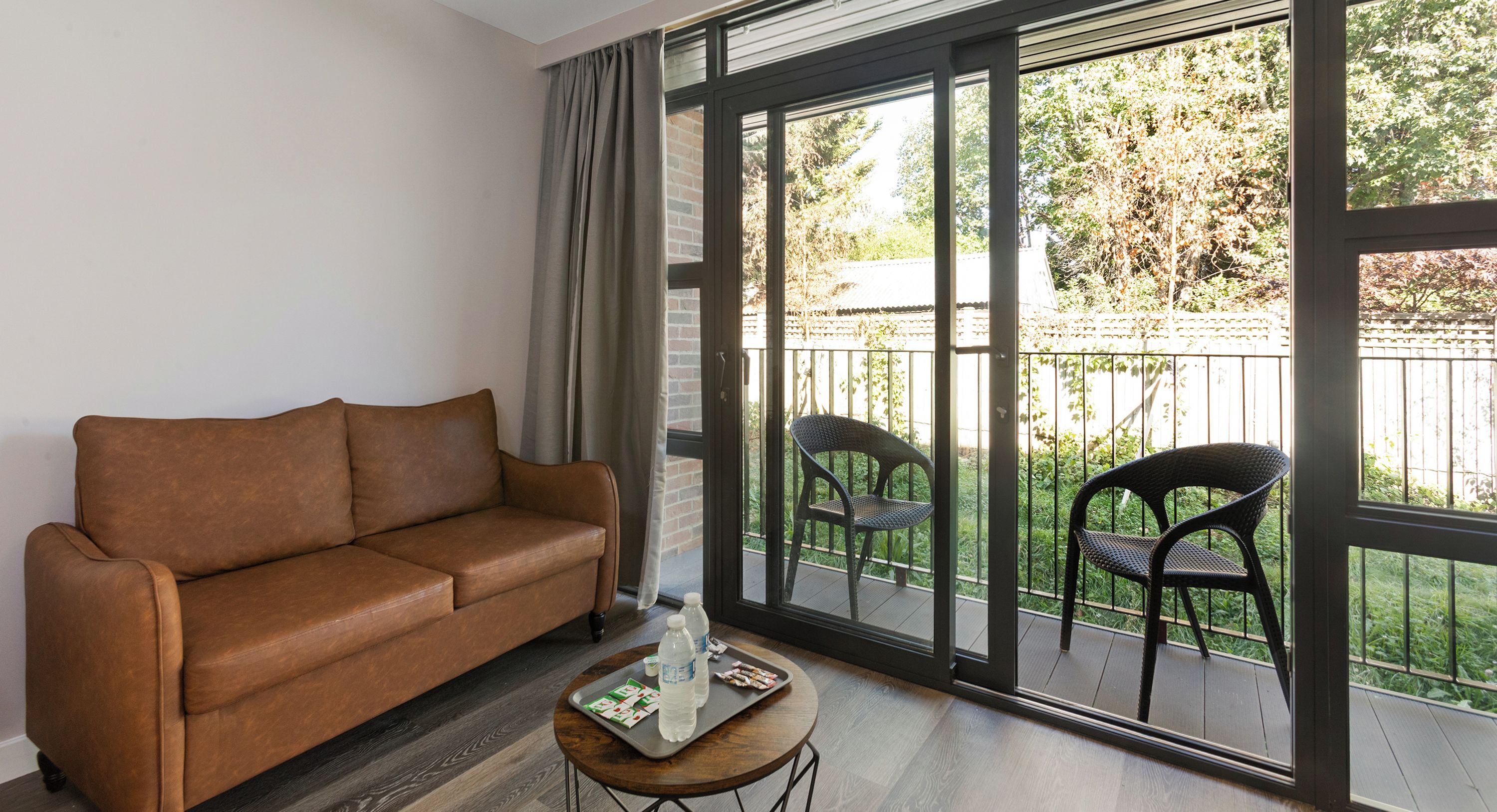 A living room with a brown sofa, a small round table with a tray, and two chairs on a balcony with a view of greenery.