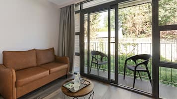 A living room with a brown sofa, a small round table with a tray, and two chairs on a balcony with a view of greenery.