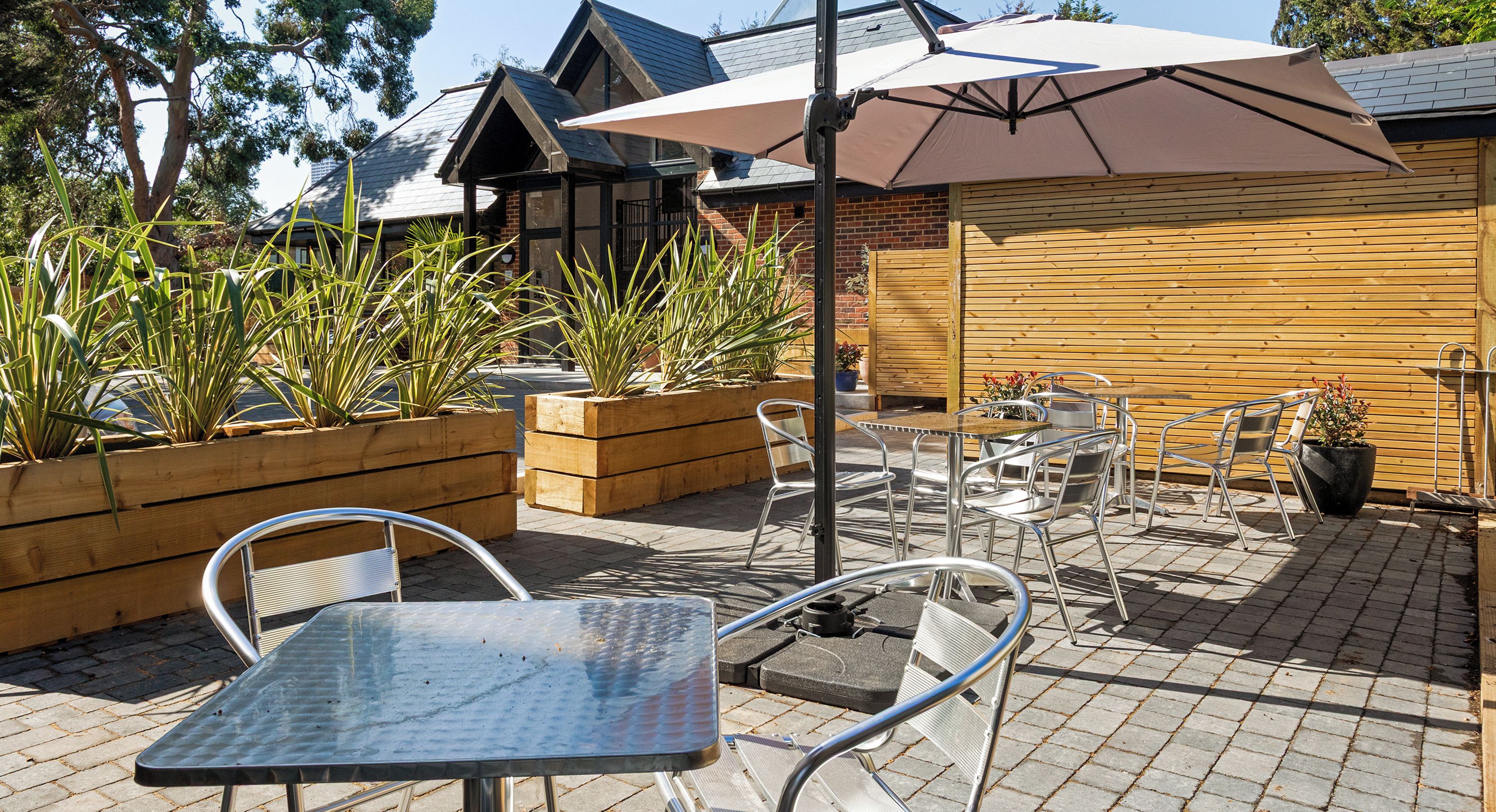 An outdoor patio with metal tables and chairs, wooden planters with plants, and a large umbrella.