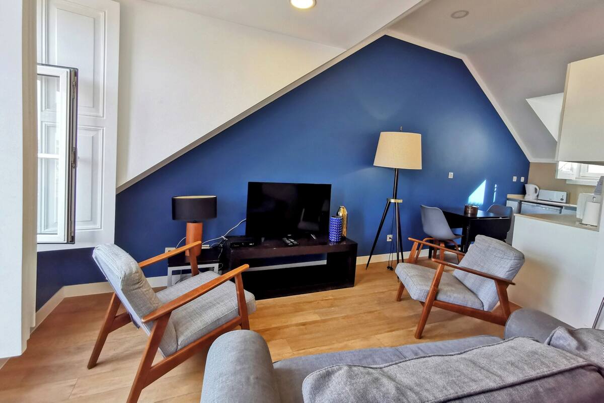 Una sala de estar moderna con televisor, dos sillones, una lámpara de pie y una cocina al fondo.