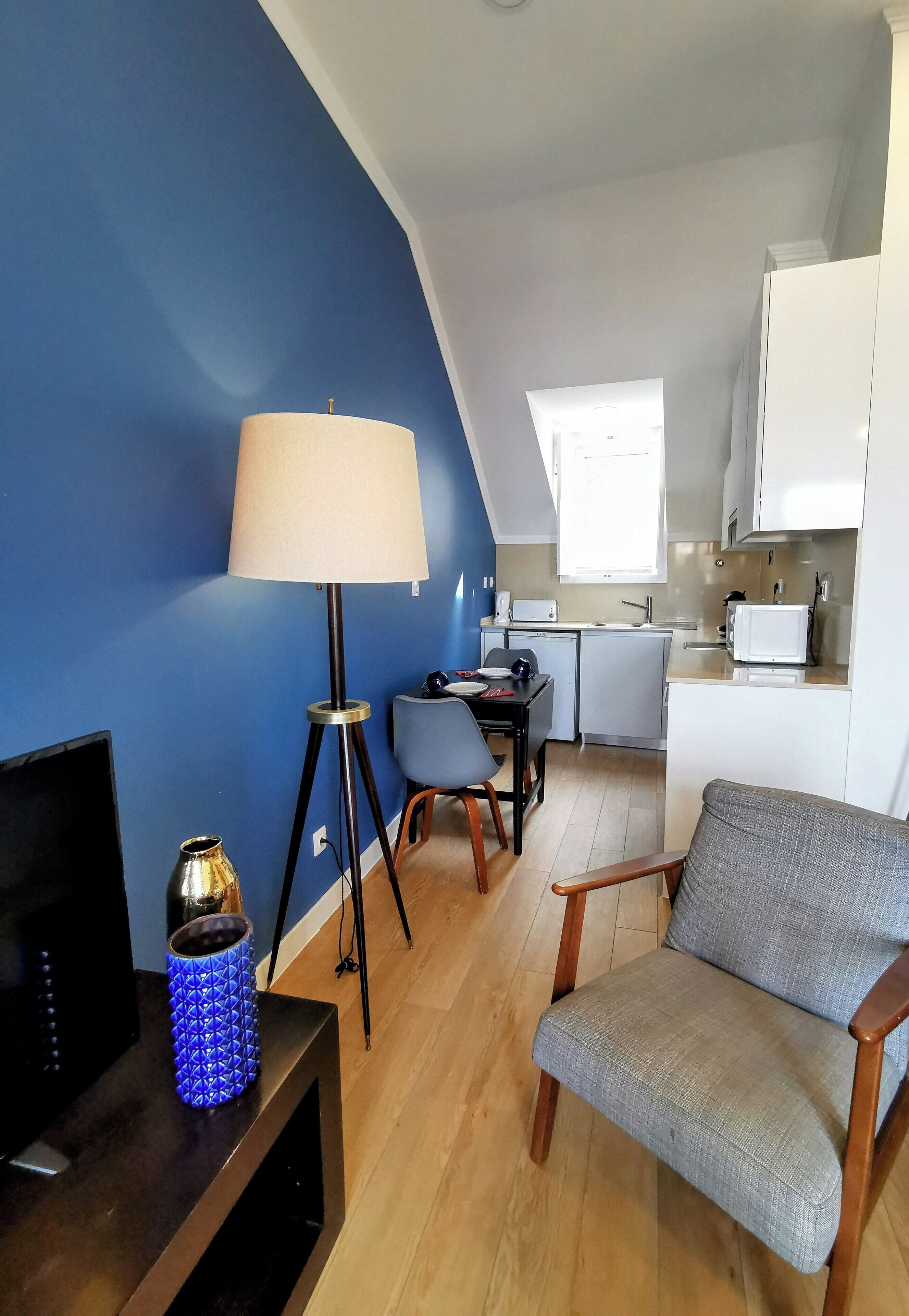 Un ambiente compatto con TV, lampada, una sedia, un tavolino e una piccola cucina a vista sullo sfondo.