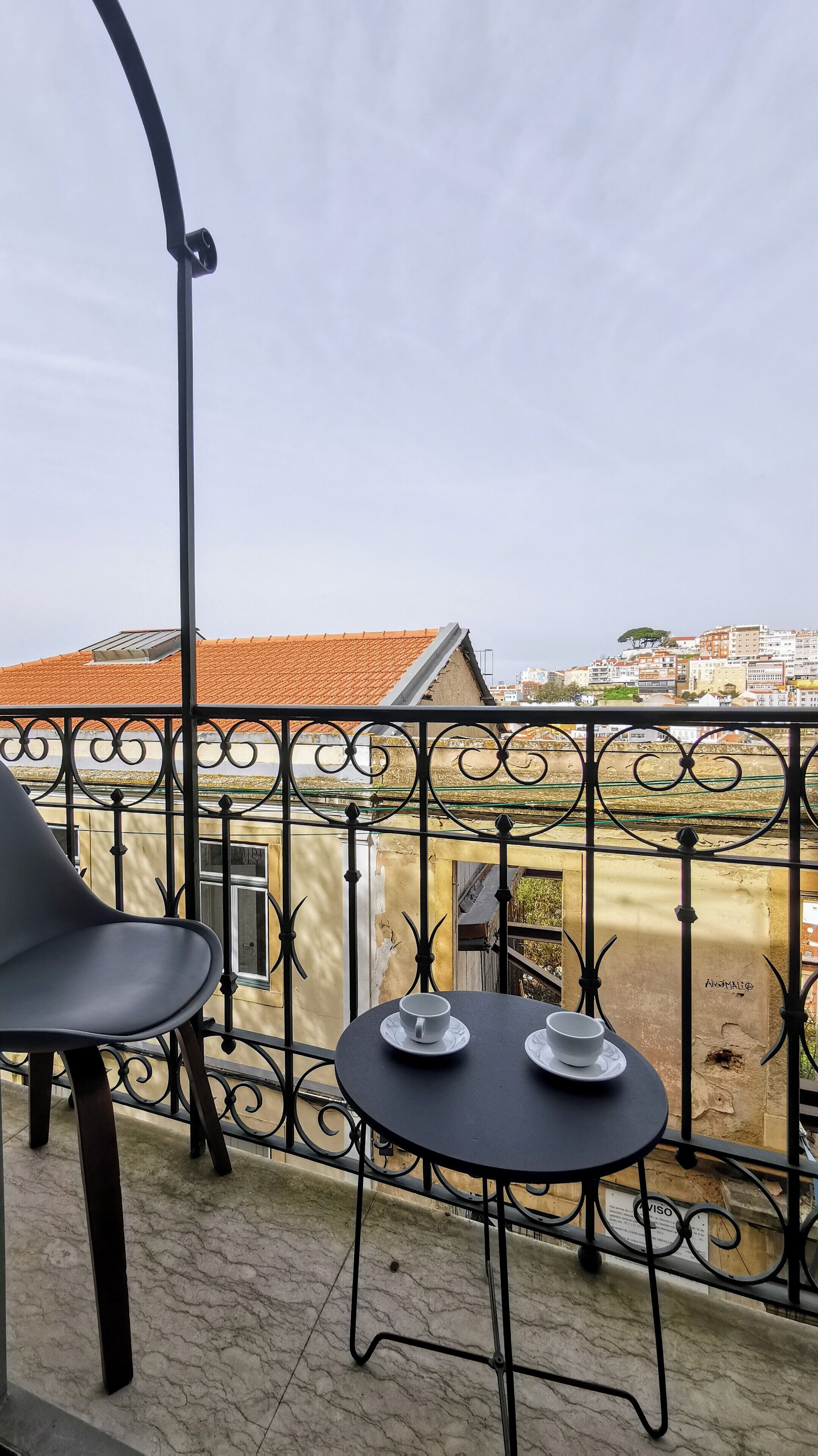 Un balcón con mesa y sillas, vista a edificios con tejados de tejas rojas y baranda de metal.