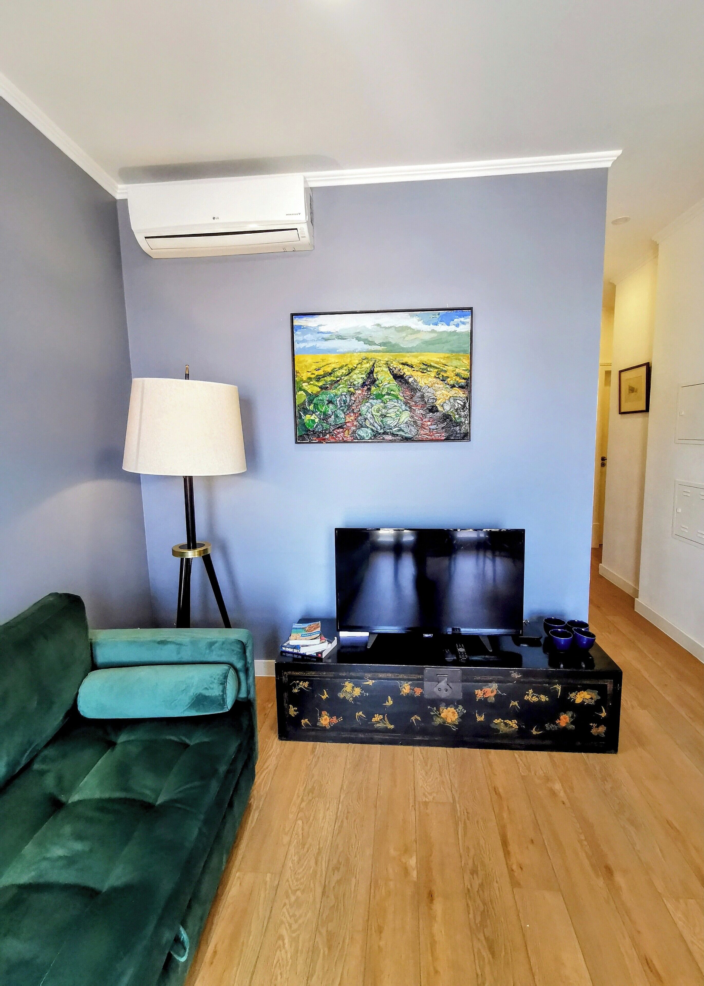 A living room with a green sofa, a floor lamp, a flat-screen TV, and a painting of a vineyard on the wall.