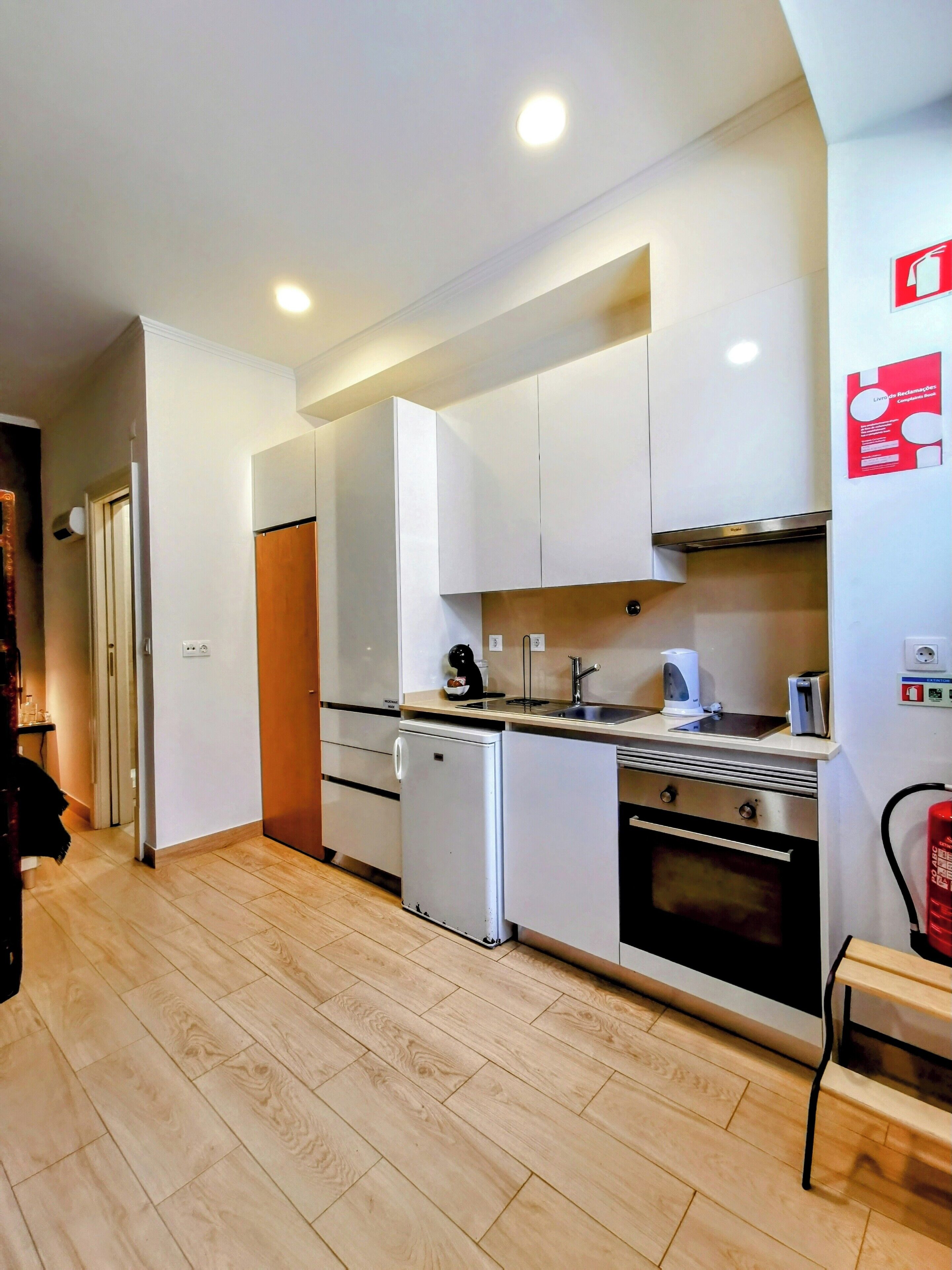 Une cuisine compacte avec des meubles blancs, un petit réfrigérateur, un lave-vaisselle et une cuisinière. On y trouve un évier avec un robinet, ainsi qu’un extincteur fixé au mur.