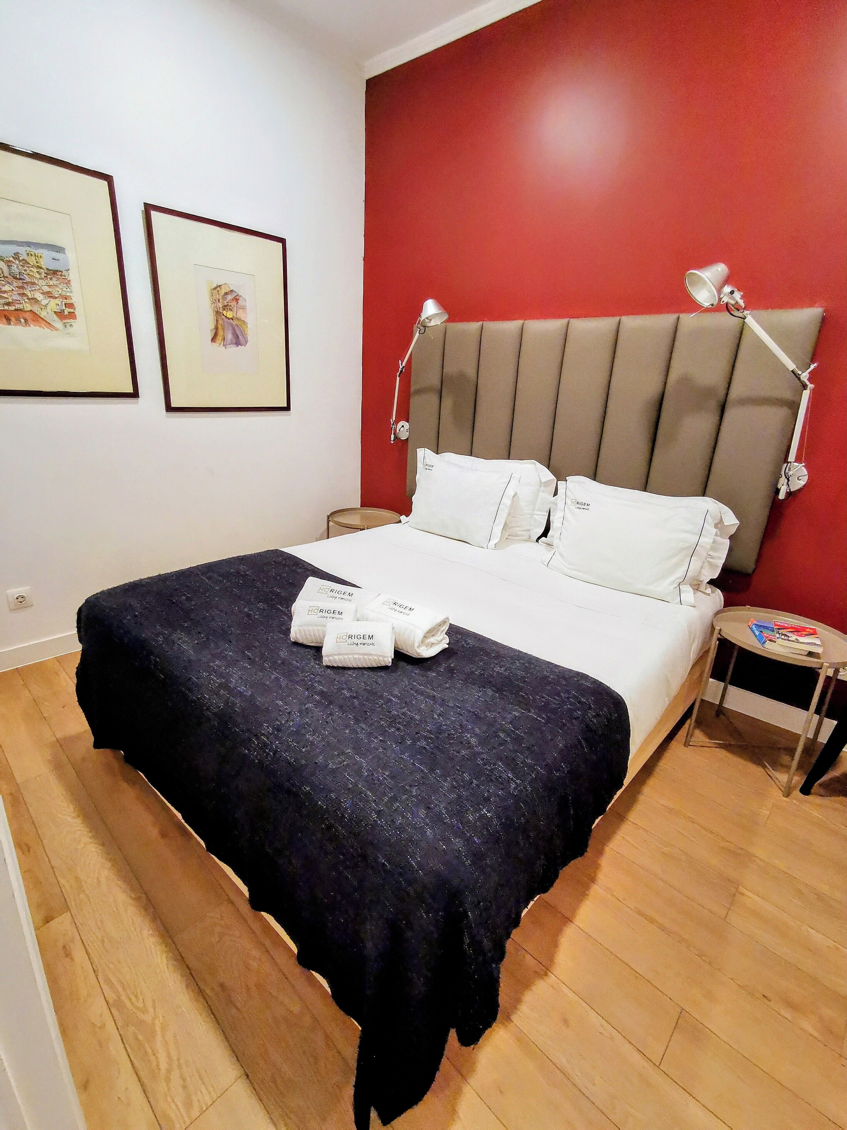Quarto de hotel com uma cama, uma mesa de cabeceira, uma parede vermelha e duas imagens emolduradas.