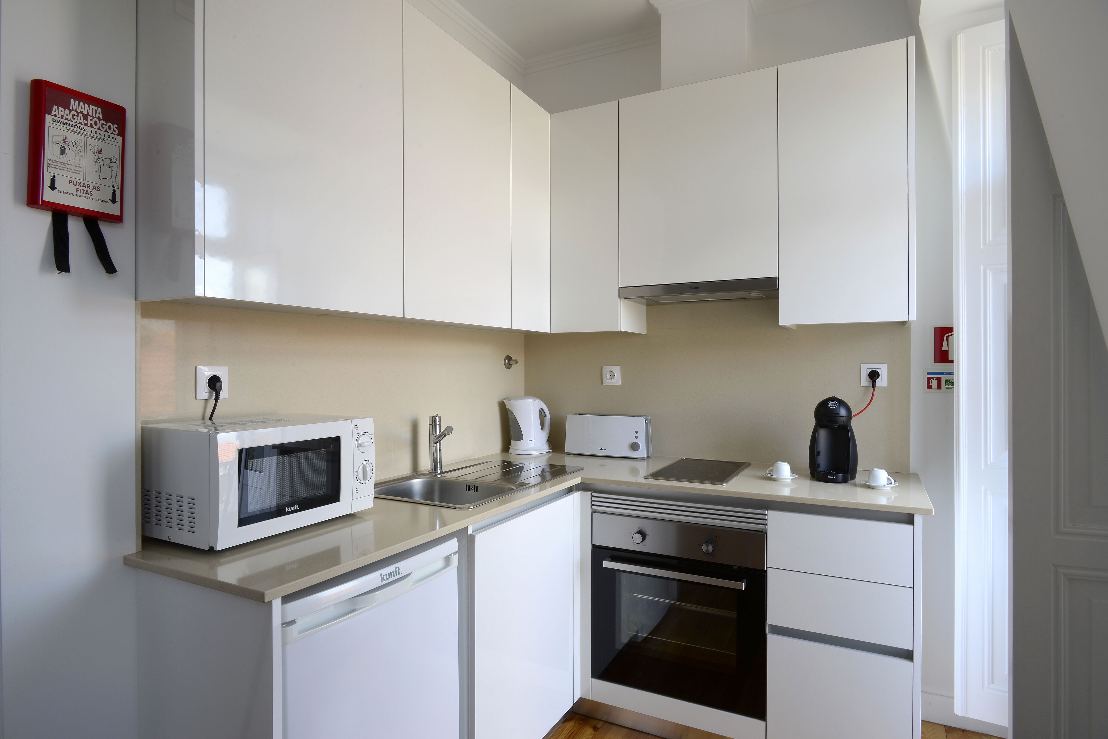 Una cocina compacta con armarios blancos, microondas, hervidor de agua, cafetera y lavavajillas.