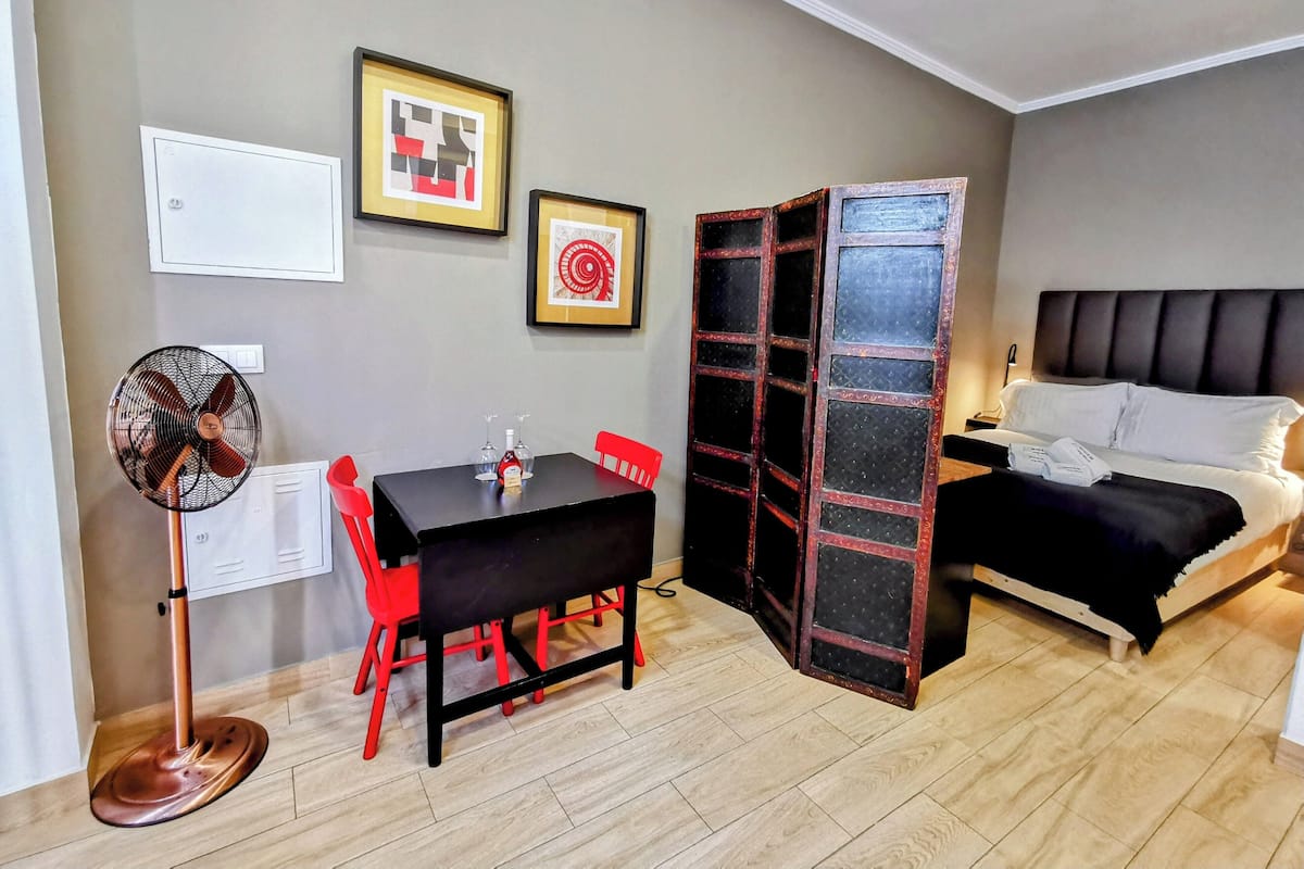 Una habitación compacta con una cama, una mesa de comedor con sillas, un ventilador de pie y dos cuadros enmarcados en la pared.