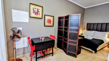 A compact room with a bed, a dining table with chairs, a standing fan, and two framed artworks on the wall.