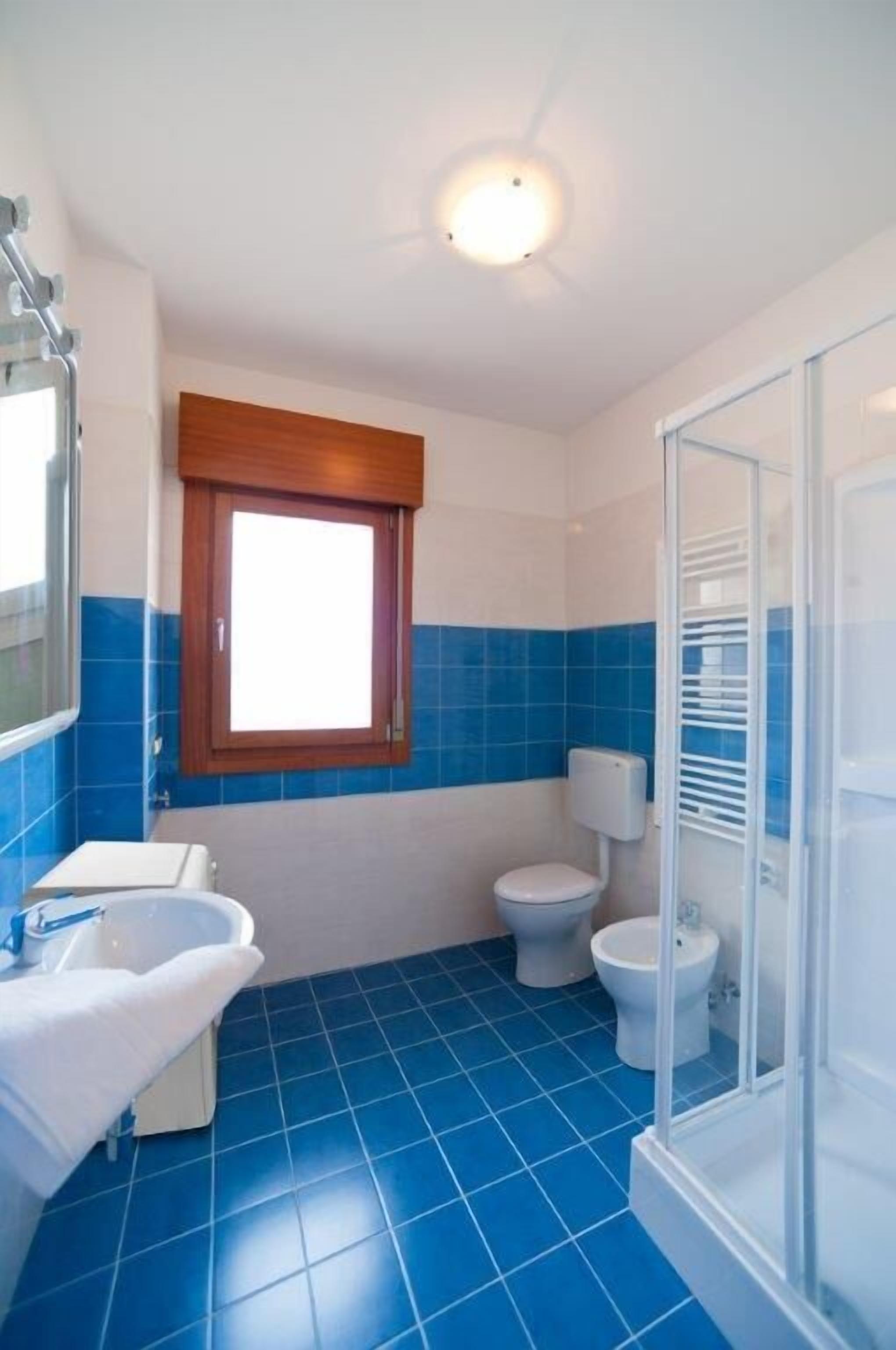 A bathroom with blue tiles, a window, a shower, and two toilets.