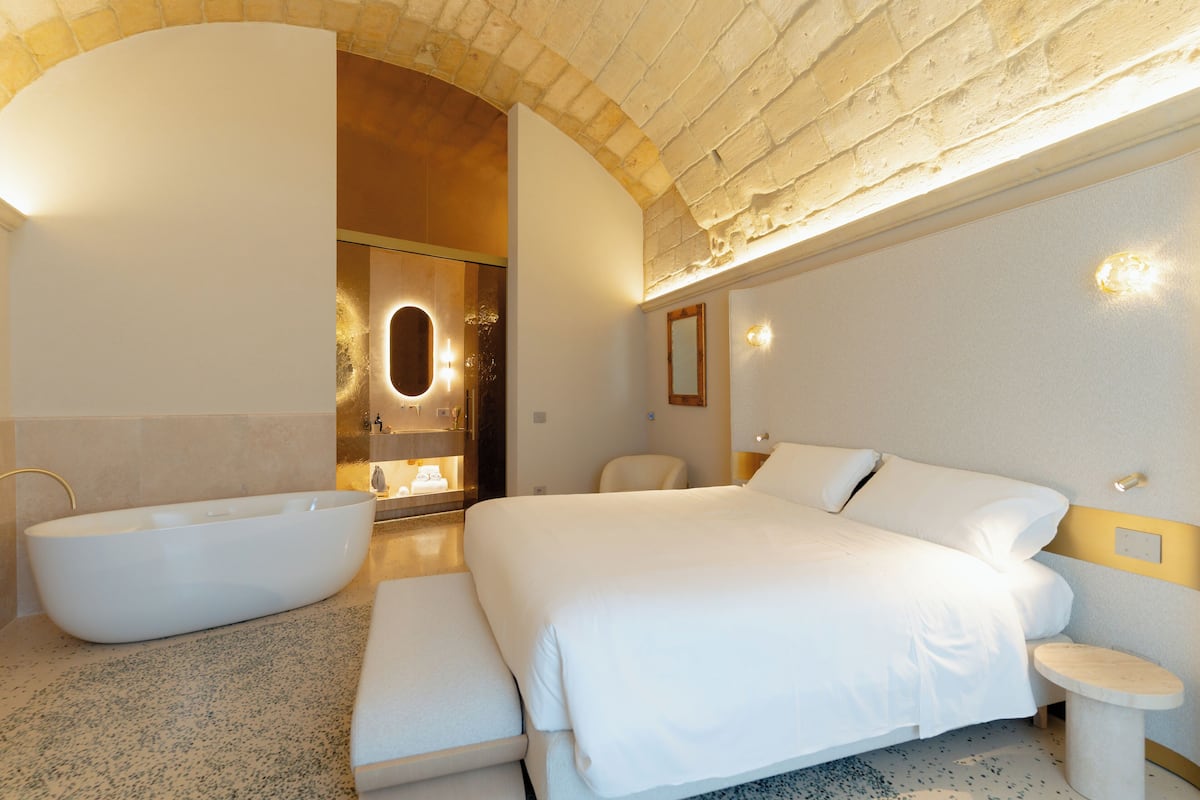 A modern hotel room with a large bed, a bathtub, and a bathroom with a mirror and lights.