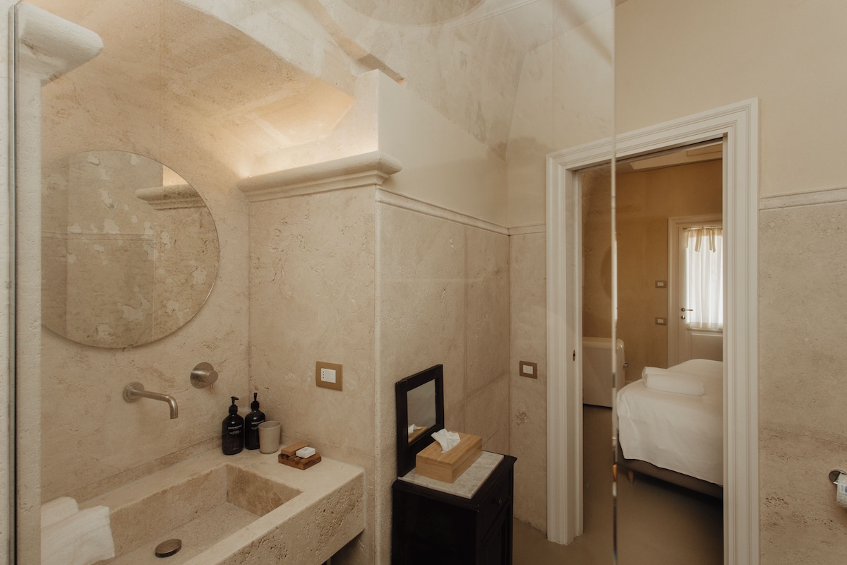 A bathroom with a marble sink, a mirror, a towel rack, and a bedside table with a box.