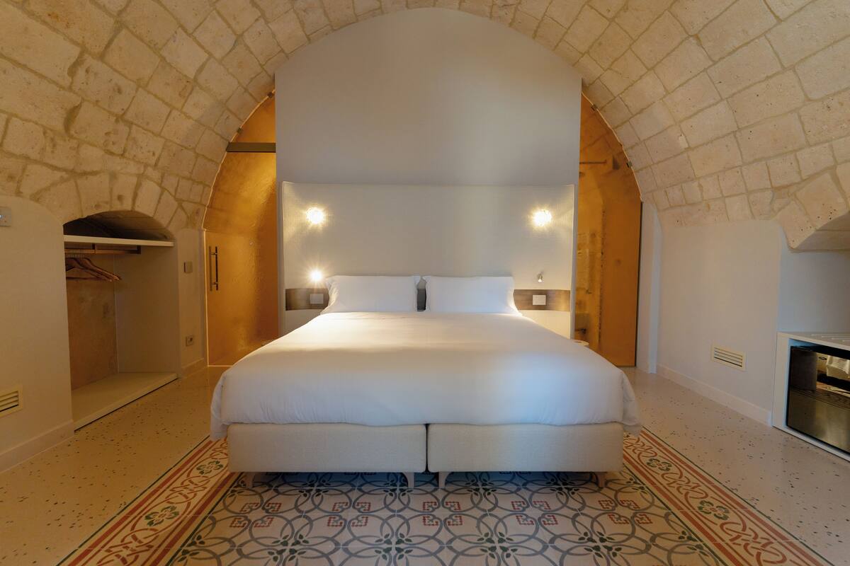 A spacious bedroom with a large bed, an arched stone ceiling, and a patterned floor.