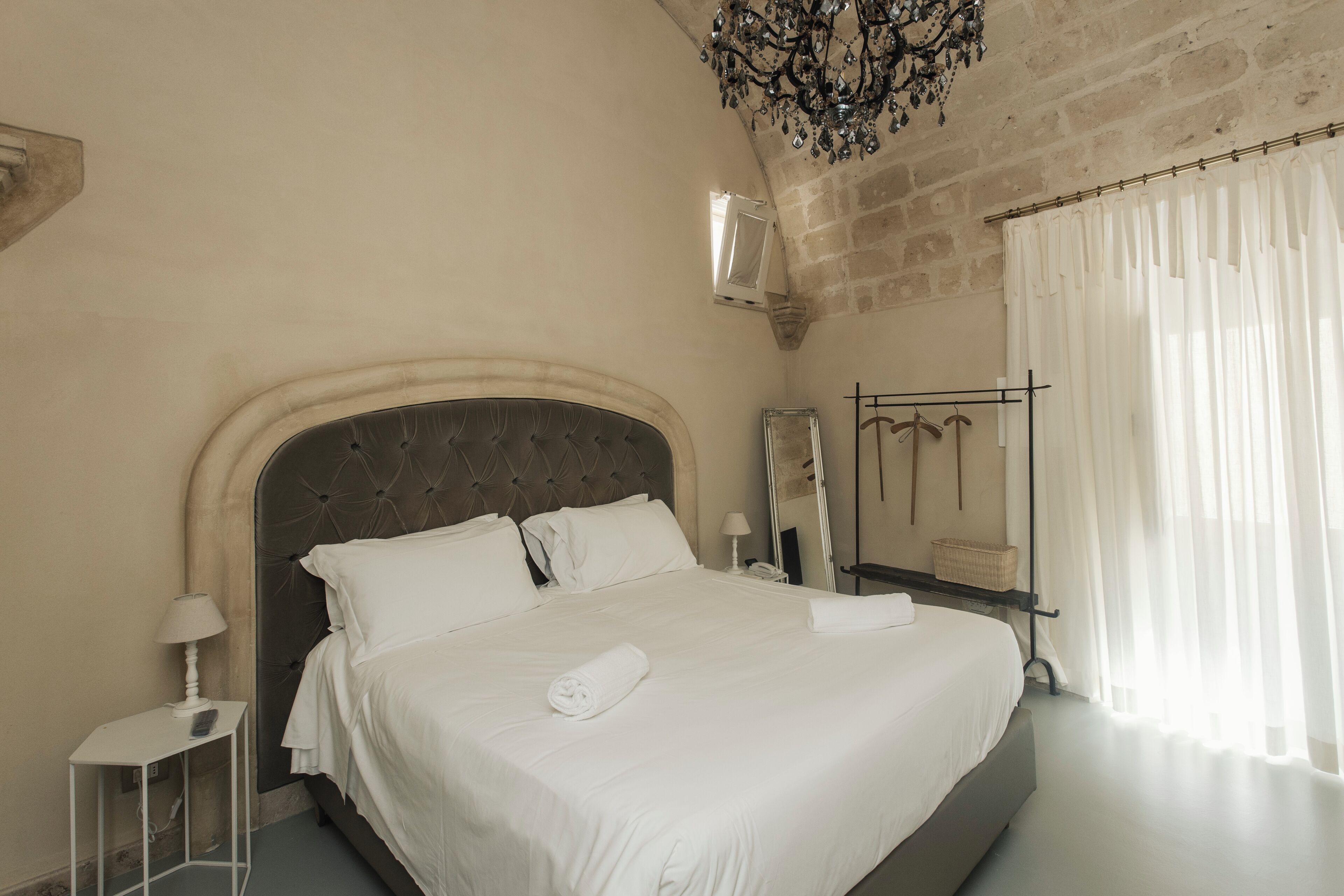 A bedroom with a large bed, a nightstand, a lamp, a mirror, and a window with sheer curtains.