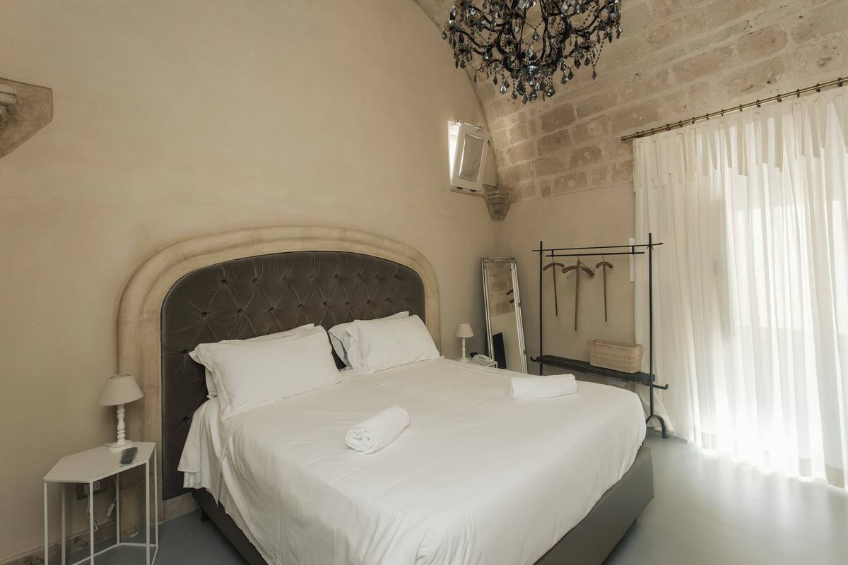 A bedroom with a large bed, a nightstand, a lamp, a mirror, and a window with sheer curtains.