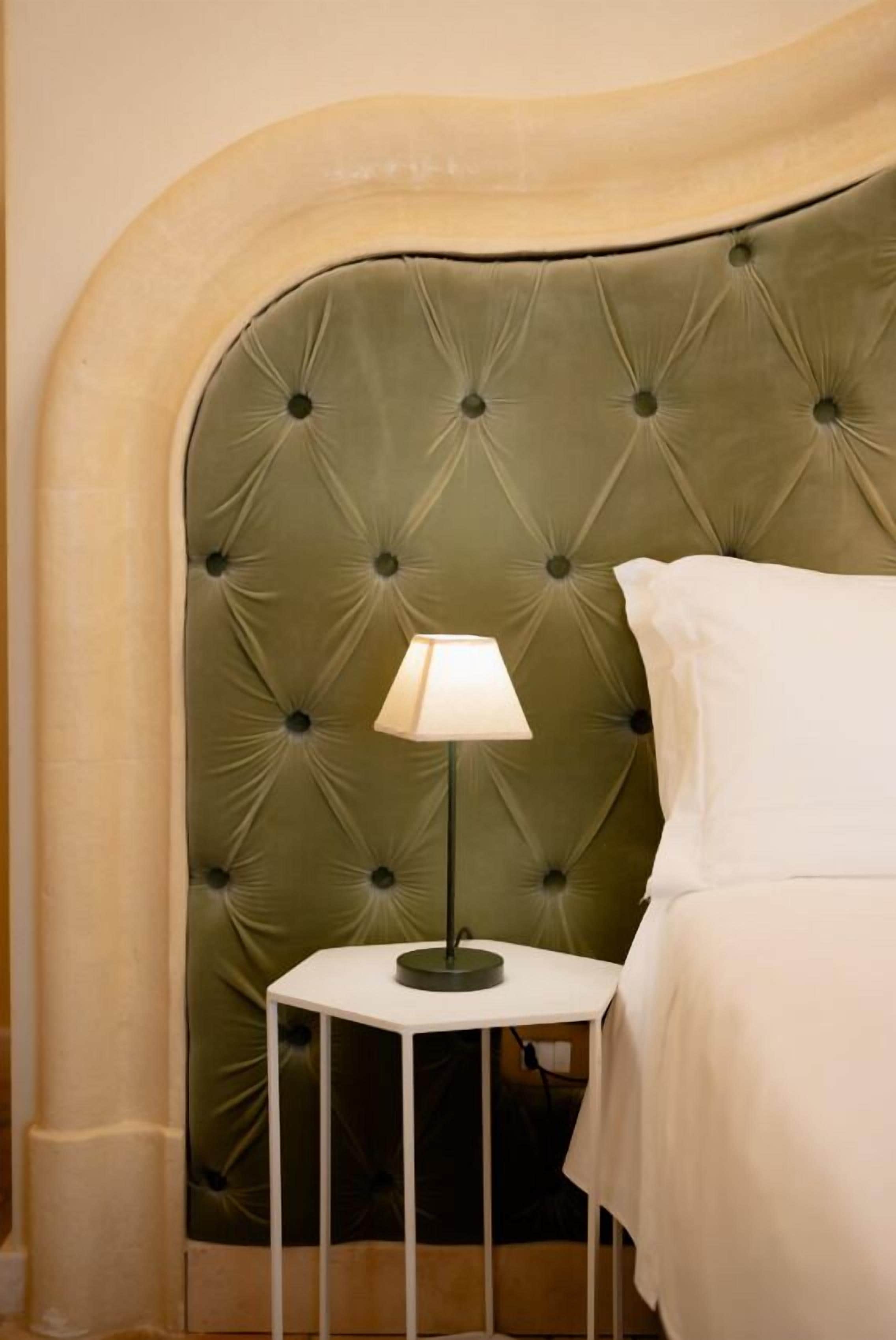 Un letto con testiera imbottita, un comodino con una lampada e un cuscino bianco.