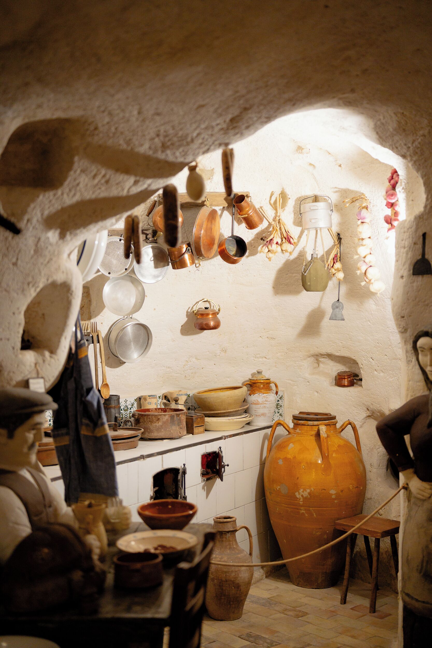 A rustic kitchen with stone walls, various cooking utensils, and a large clay pot.