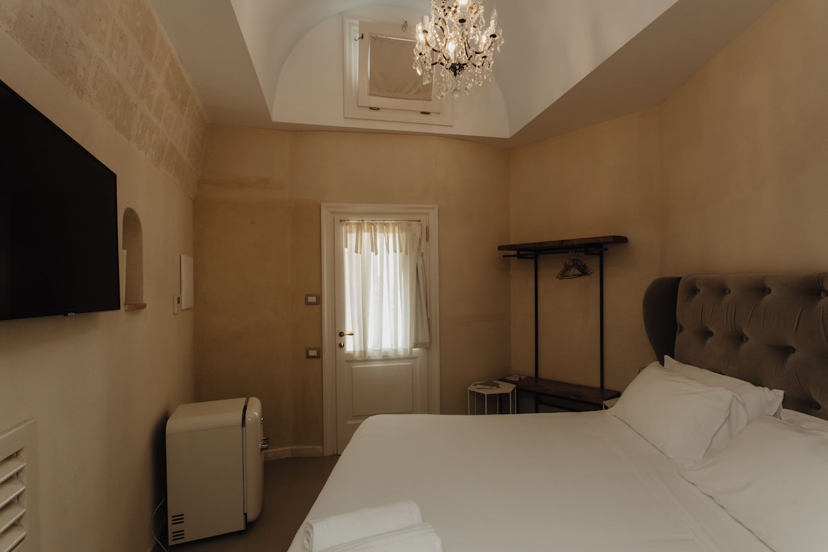 A hotel room with a bed, a television, a chandelier, a door, and a small table.