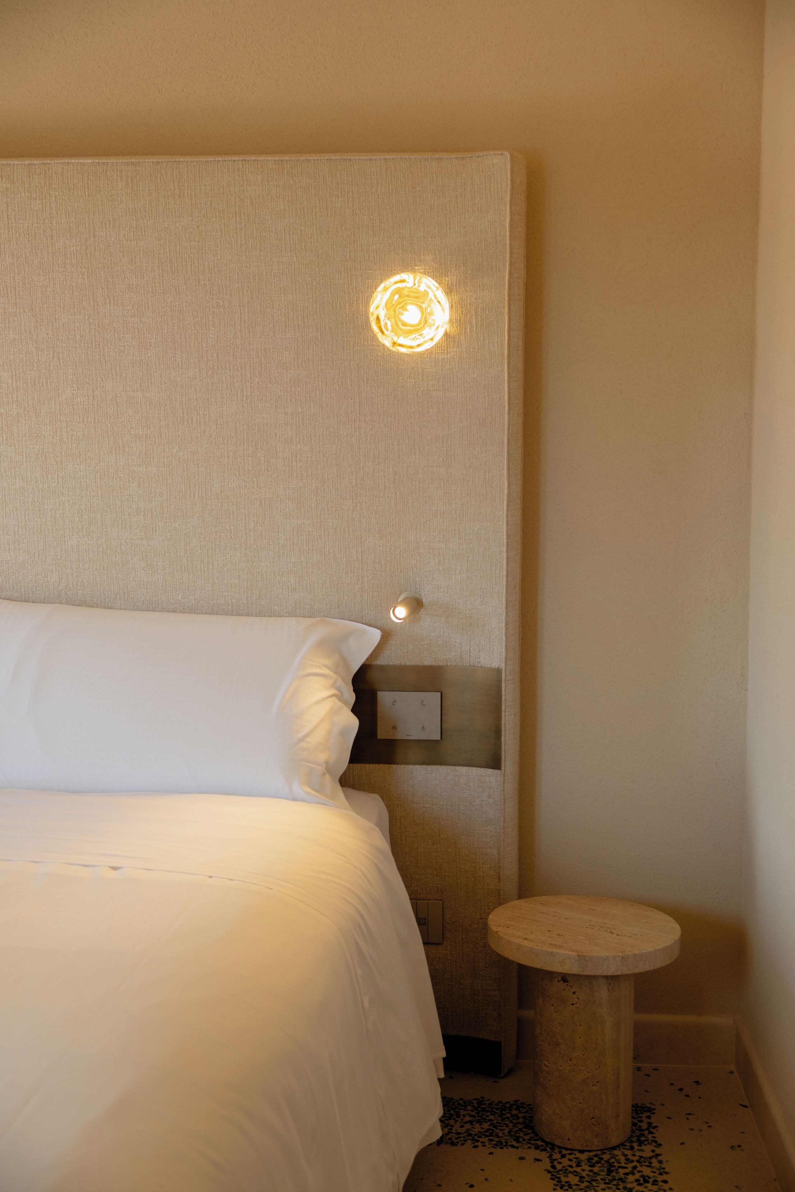 A neatly made bed with white linens, a small wooden stool, and a wall-mounted light fixture.