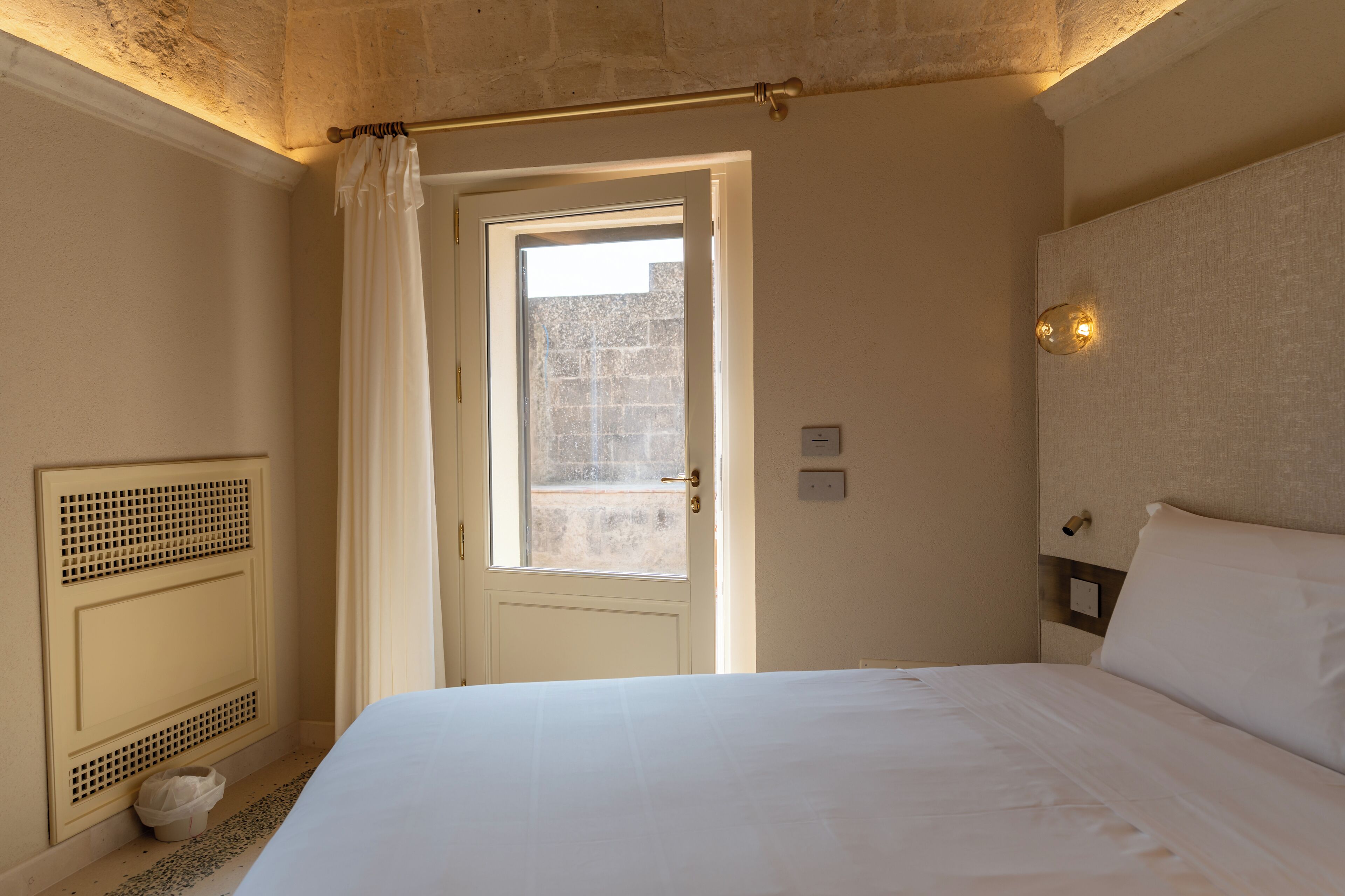A hotel room with a bed, a window with curtains, a radiator, and a wall-mounted light fixture.