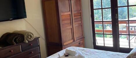 A bedroom with a bed, a wooden wardrobe, a television mounted on the wall, and a window with a view of greenery.