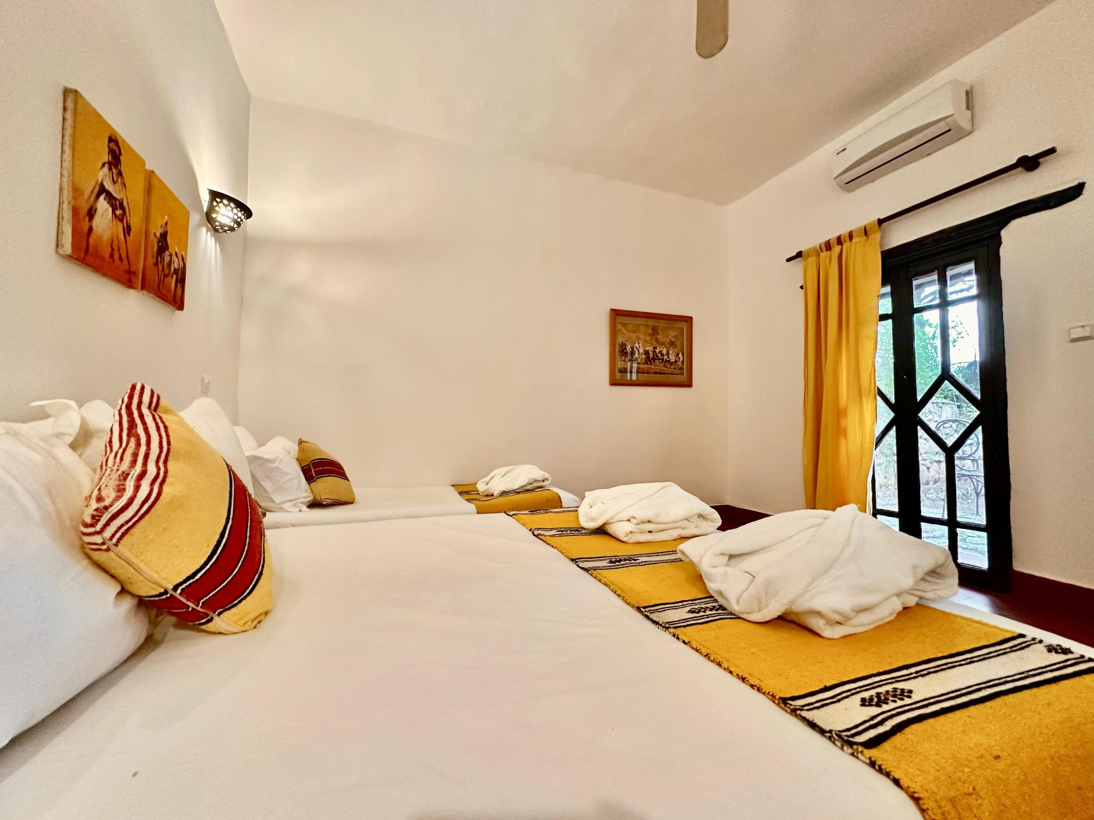 A hotel room with two beds, a window with curtains, an air conditioning unit, and artwork on the walls.