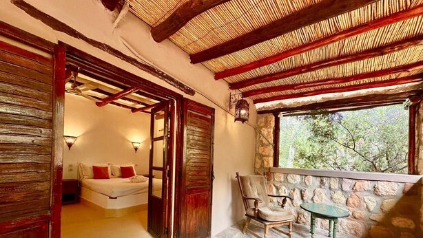 A room with a stone floor, a bed with red pillows, a wooden door, a stone wall, a window with a view of greenery, and a small table with a chair. - La Bergerie (Ouirgane)