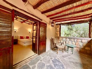 Quarto com piso de pedra, cama com almofadas vermelhas, porta de madeira, parede de pedra, janela com vista para o verde e uma pequena mesa com cadeira.