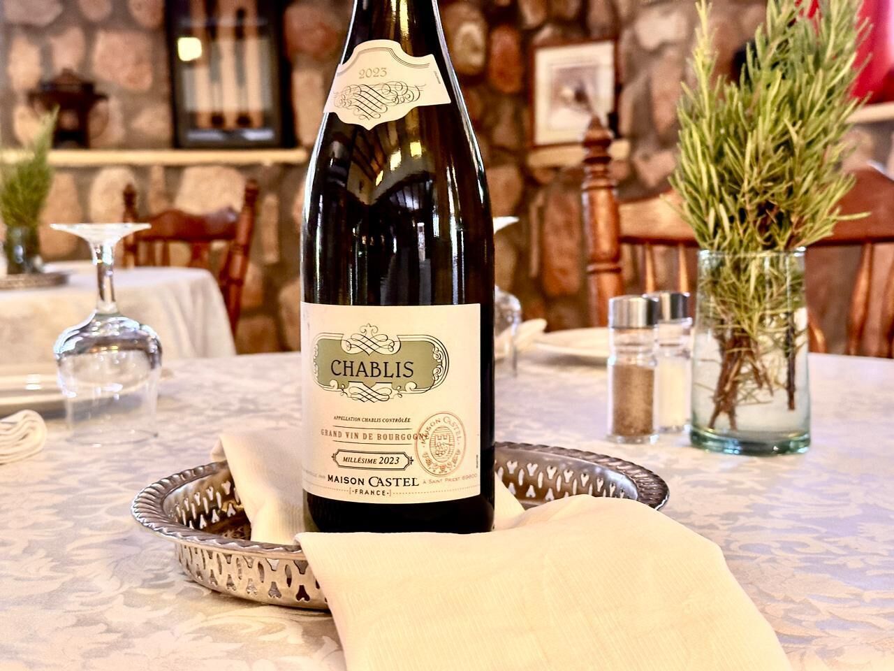 A bottle of Chablis wine with a label, placed on a table with a napkin, a glass, and a decorative vase in the background.