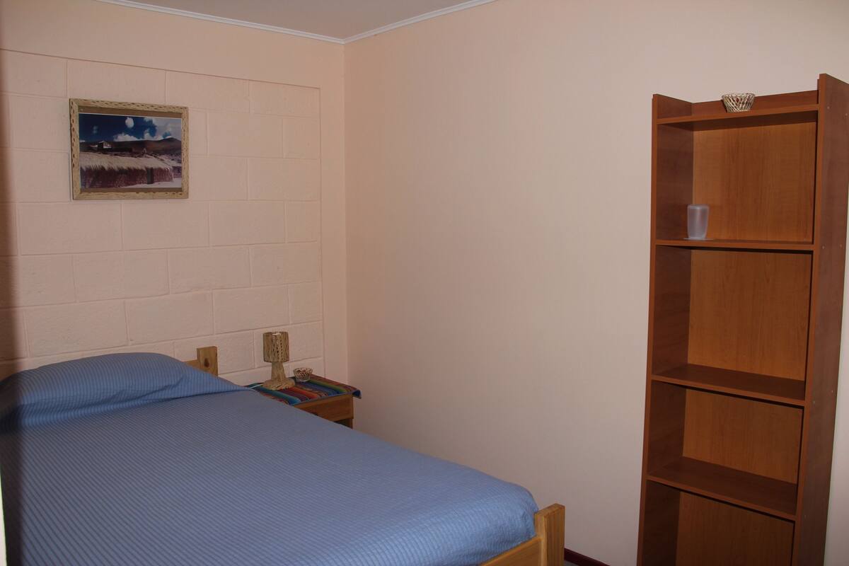 A bedroom with a single bed, a wooden bookshelf, a small table with a lamp, and a framed picture on the wall.