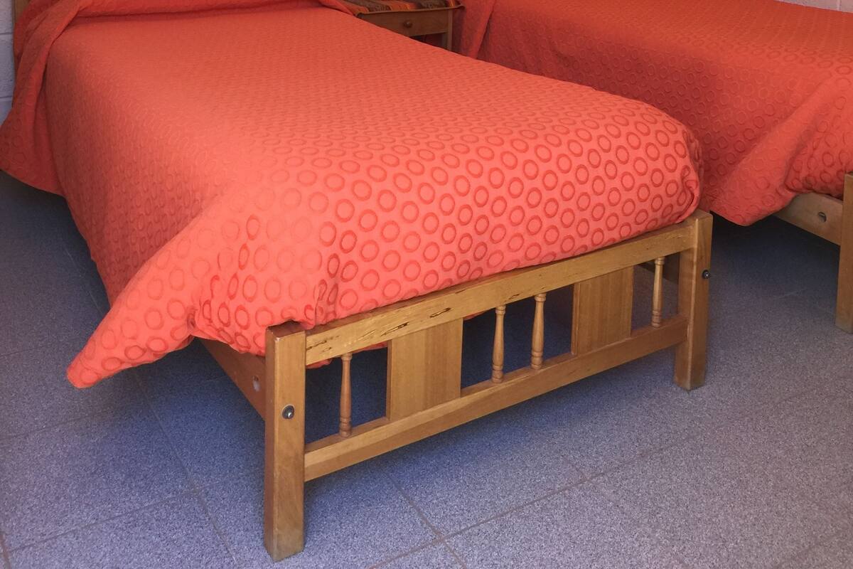 A room with two wooden beds, each with a red patterned bedspread. There is a small wooden nightstand with a lamp between the beds. The walls are white brick, and there is a framed picture on the wall above the beds.