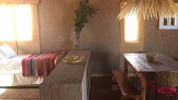 Una habitación rústica con una isla de hormigón, un banco de madera, una silla de fibra natural, una mesa con una cesta y una planta.