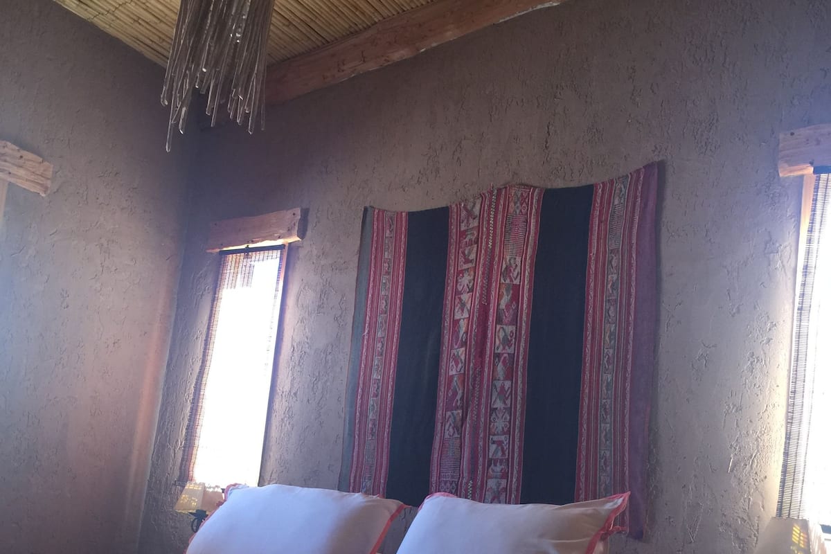 A bed with white linens and a striped blanket, a wooden ceiling, and a window with patterned curtains.