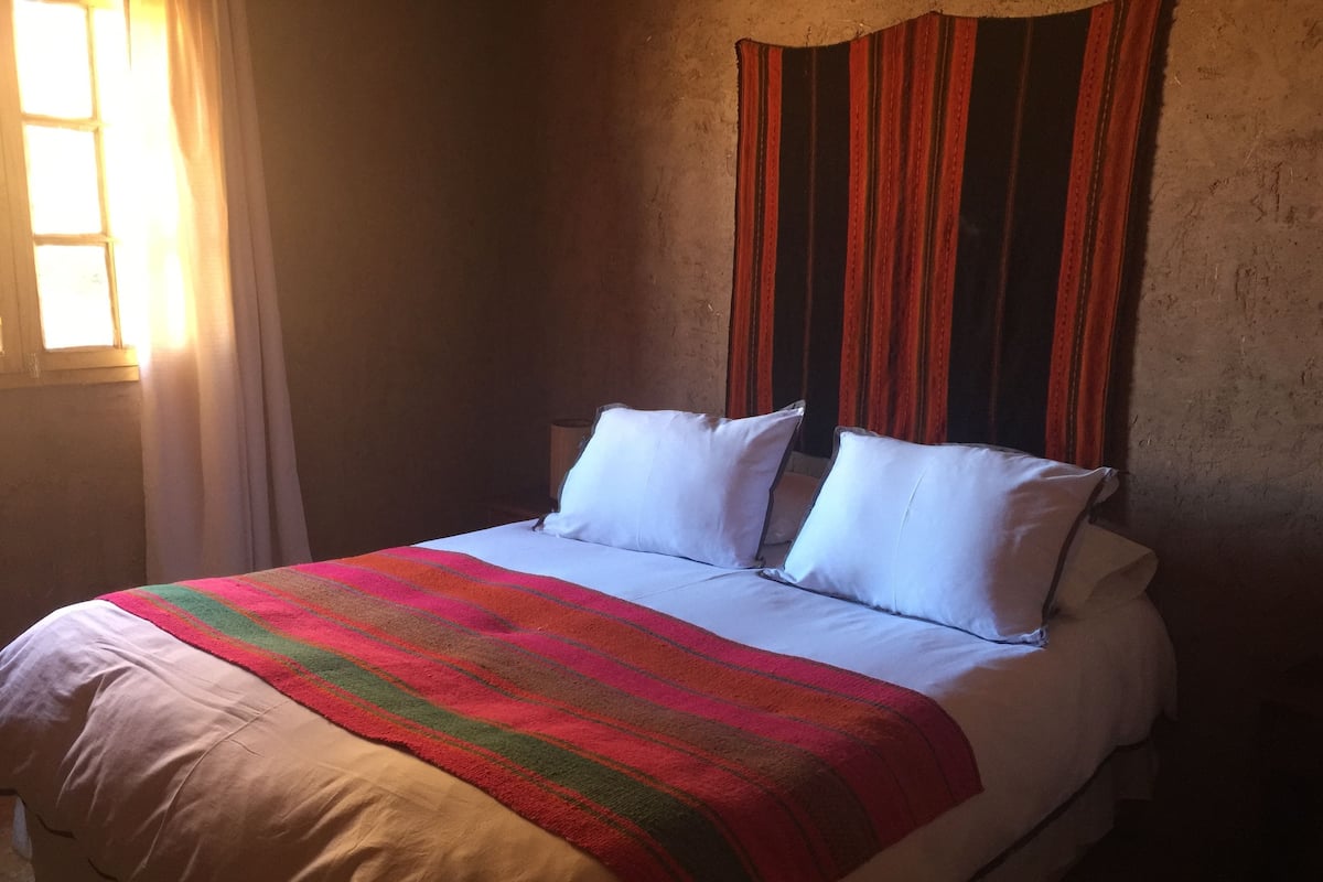 A bed with white linens and a colorful striped blanket, a wooden headboard, and a window with curtains.