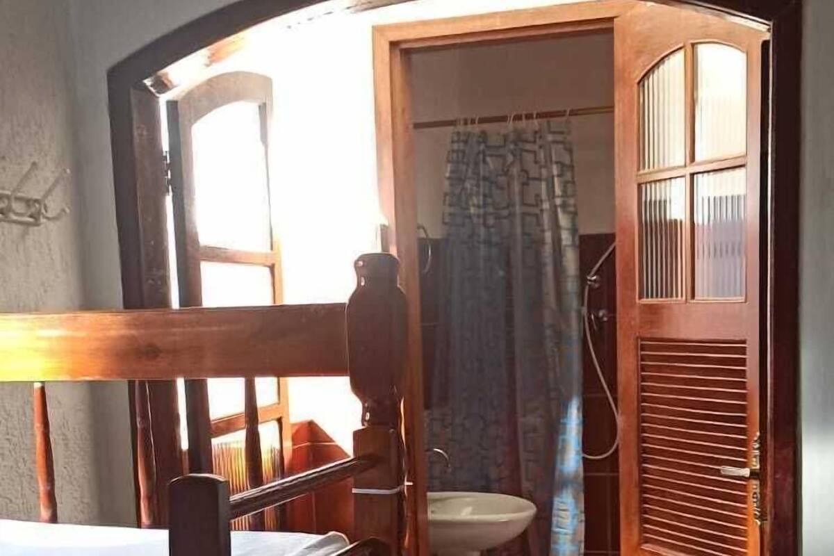 A bedroom with a wooden bed, a window, and an open door leading to a bathroom with a sink and shower curtain.