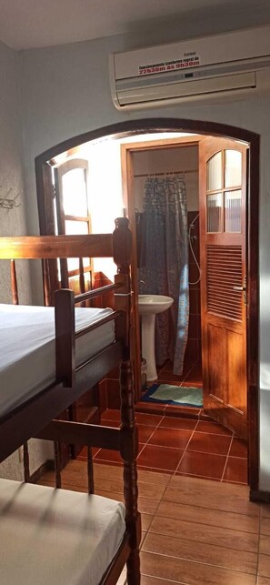 A bedroom with a wooden bed, a window, and an open door leading to a bathroom with a sink and shower curtain. - 365 Hostel (Angra dos Reis)