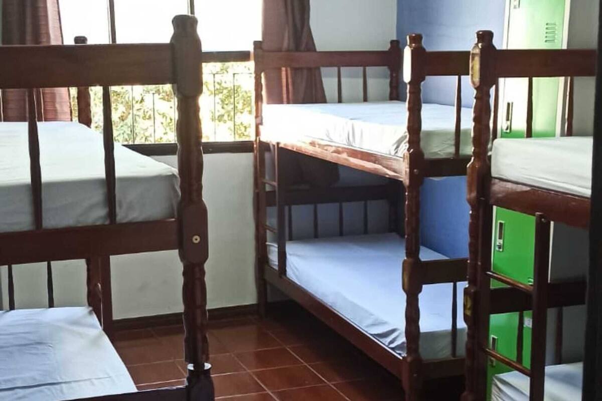 A dormitory room with bunk beds, a window with curtains, an air conditioning unit, and a green storage cabinet.