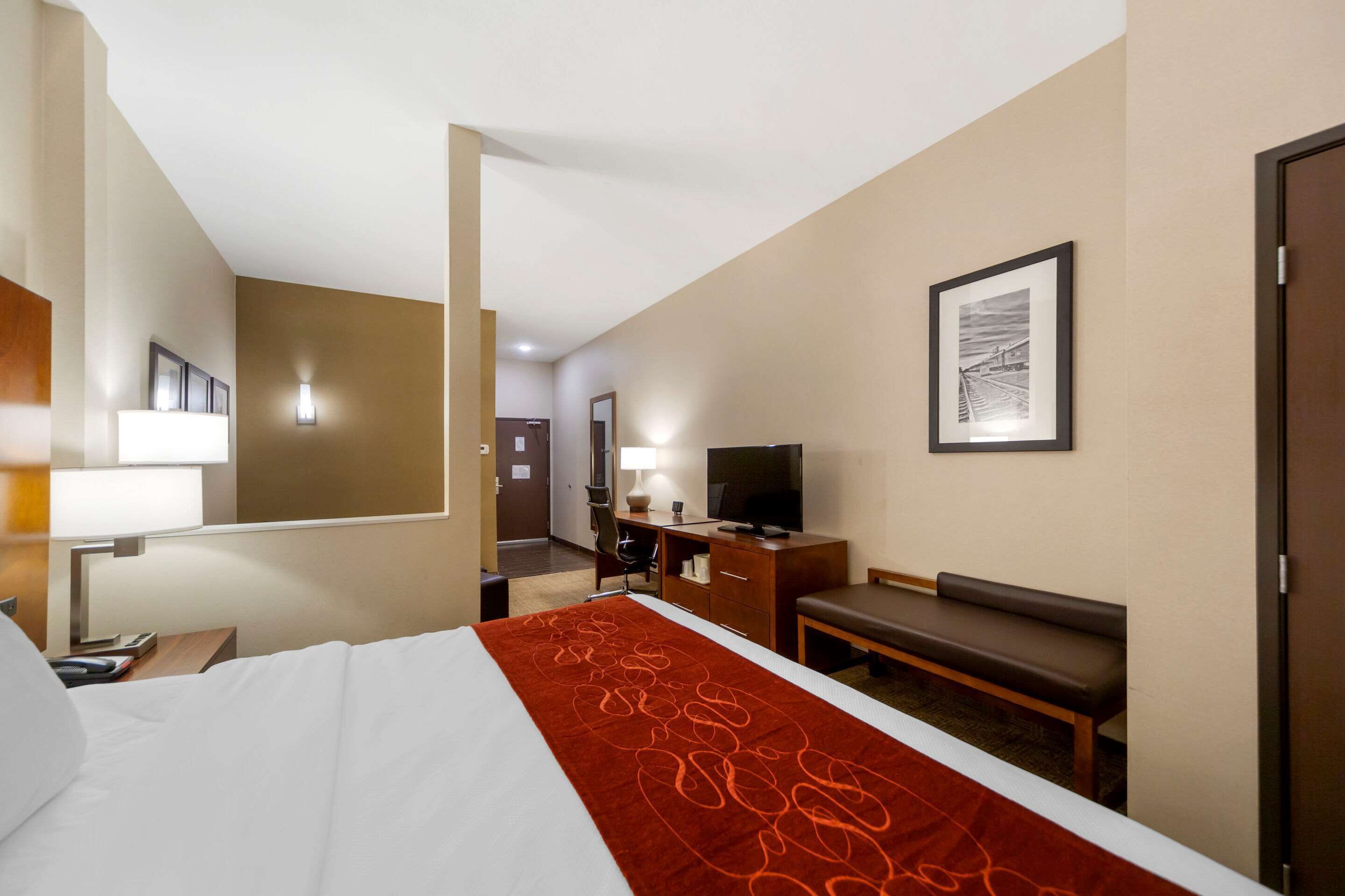Ein Hotelzimmer mit einem Bett, einem Schreibtisch mit Computer, einem Fernseher, einer Bank und einem gerahmten Bild an der Wand.