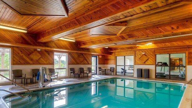 Une piscine intérieure avec des poutres en bois au plafond, un espace détente avec des fauteuils et du matériel d’exercice.