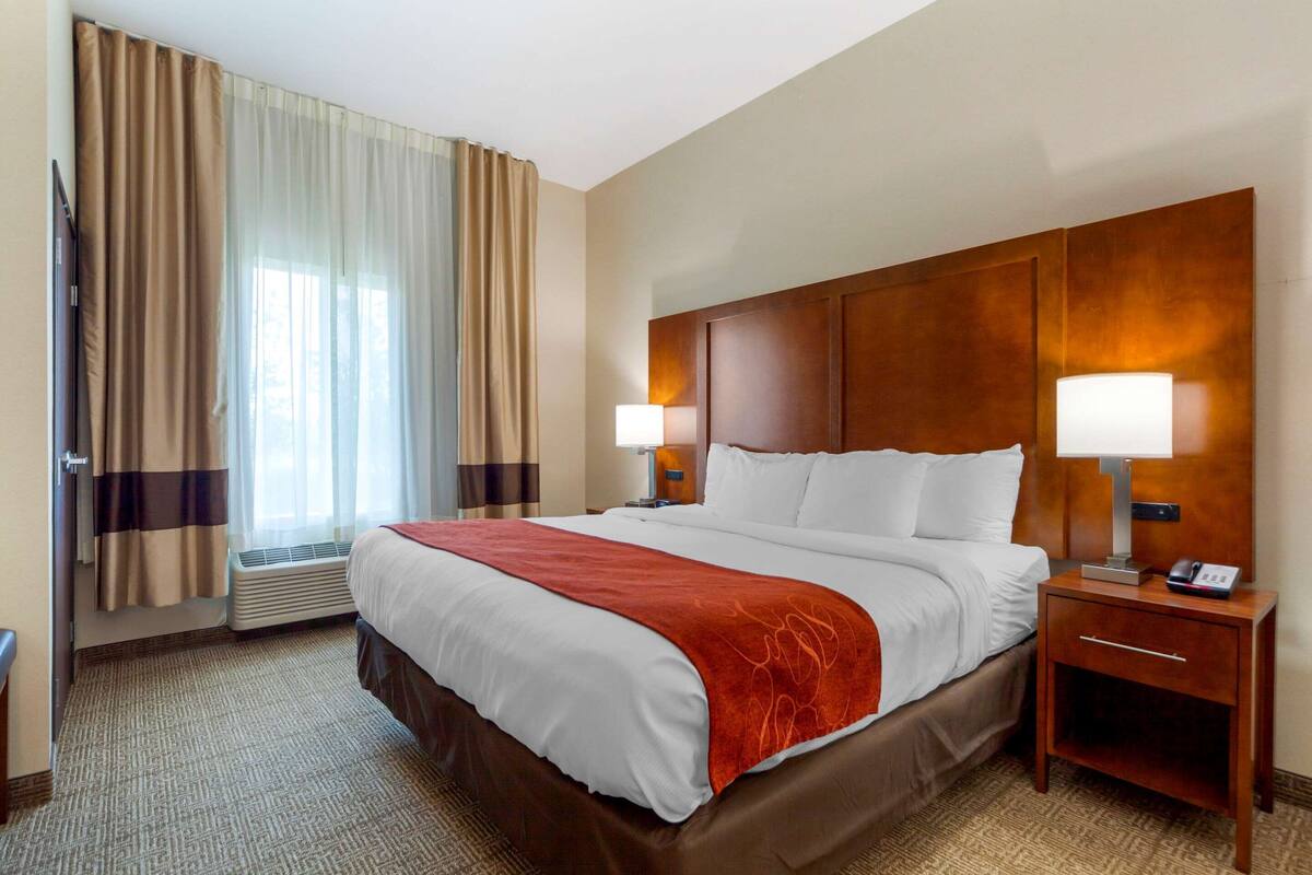 Ein Hotelzimmer mit einem großen Bett, zwei Nachttischlampen, einem hölzernen Kopfteil, einem Nachttisch mit Telefon und einem Fenster mit Vorhängen.